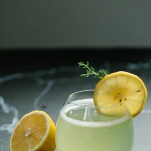 Close-up of a citrus beverage in a glass, garnished with a lemon slice and fresh herb on a dark surface.
